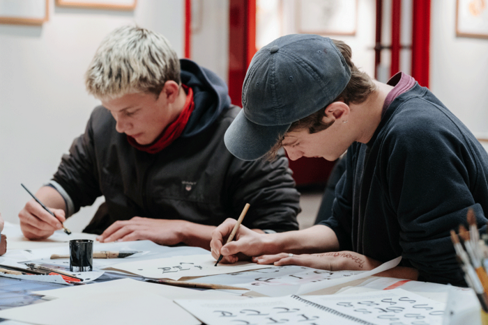 Zwei Jugendliche sitzen an einem Tisch und zeichnen mit Tusche Schriftzeichen.