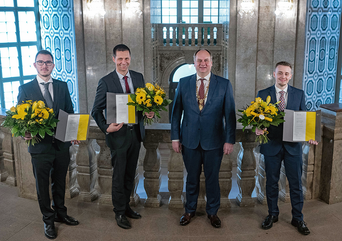 Foto aus der Einganghalle im Neuen rathaus Dresden: Der Oberbürgermeister Dirk Hilbert steht mit drei Herren zusammen, welche Blumensträuße und Urkunden in den Händen halten.