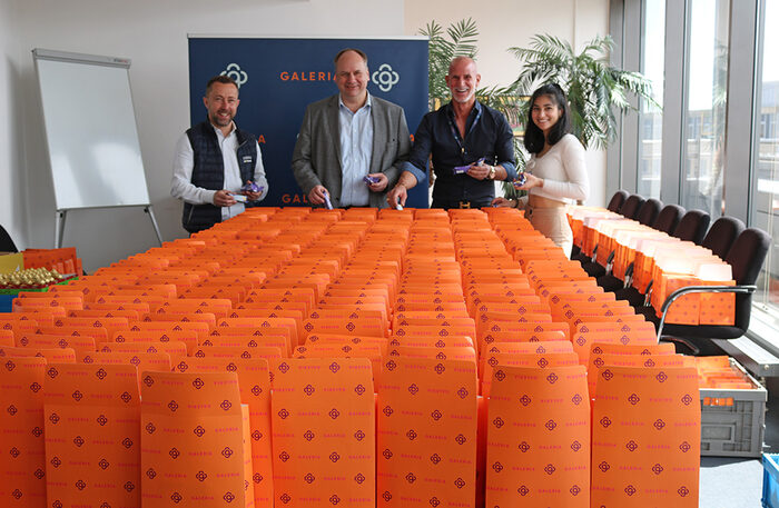 Foto von einer großen Zahl orangefarbener Geschenktüten auf einem Tisch. Dahinter stehen vier Personen.