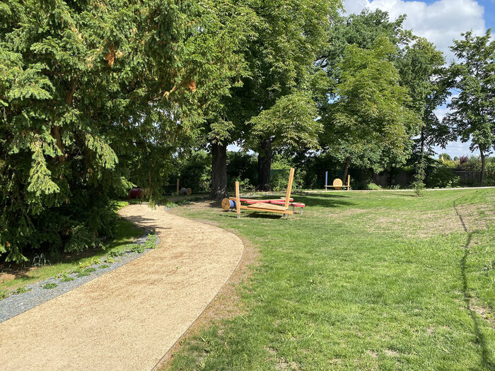 Wiese mit kleinem Spielplatz, auf der linken Seite stehen Bäume, dazwischen schlängelt sich ein kleiner Weg.