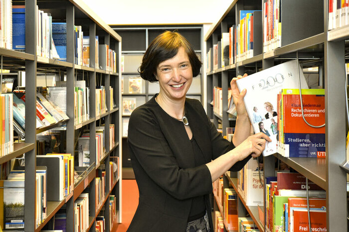 Frau mit dunklen Haaren und schwarzem Oberteil steht zwischen zwei Bücherregalen und zieht das Buch "200 Frauen" aus dem Regal. Sie lächelt in die Kamera.