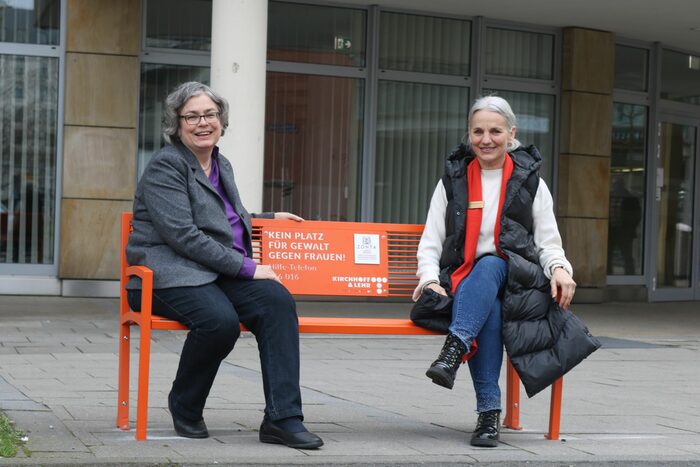 Zwei Frauen sitzen auf einer orangenen Bank vor einem Gebäude