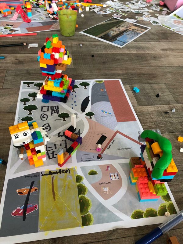 Aus Lego gebauter Baum, Katze und Haus, auf dem noch eine grüne Knete liegt stehen auf einem mit einer Landschaft bedruckten Blatt Papier. Auf diesem sind Bäume eingezeichnet und Autos durchgestrichen