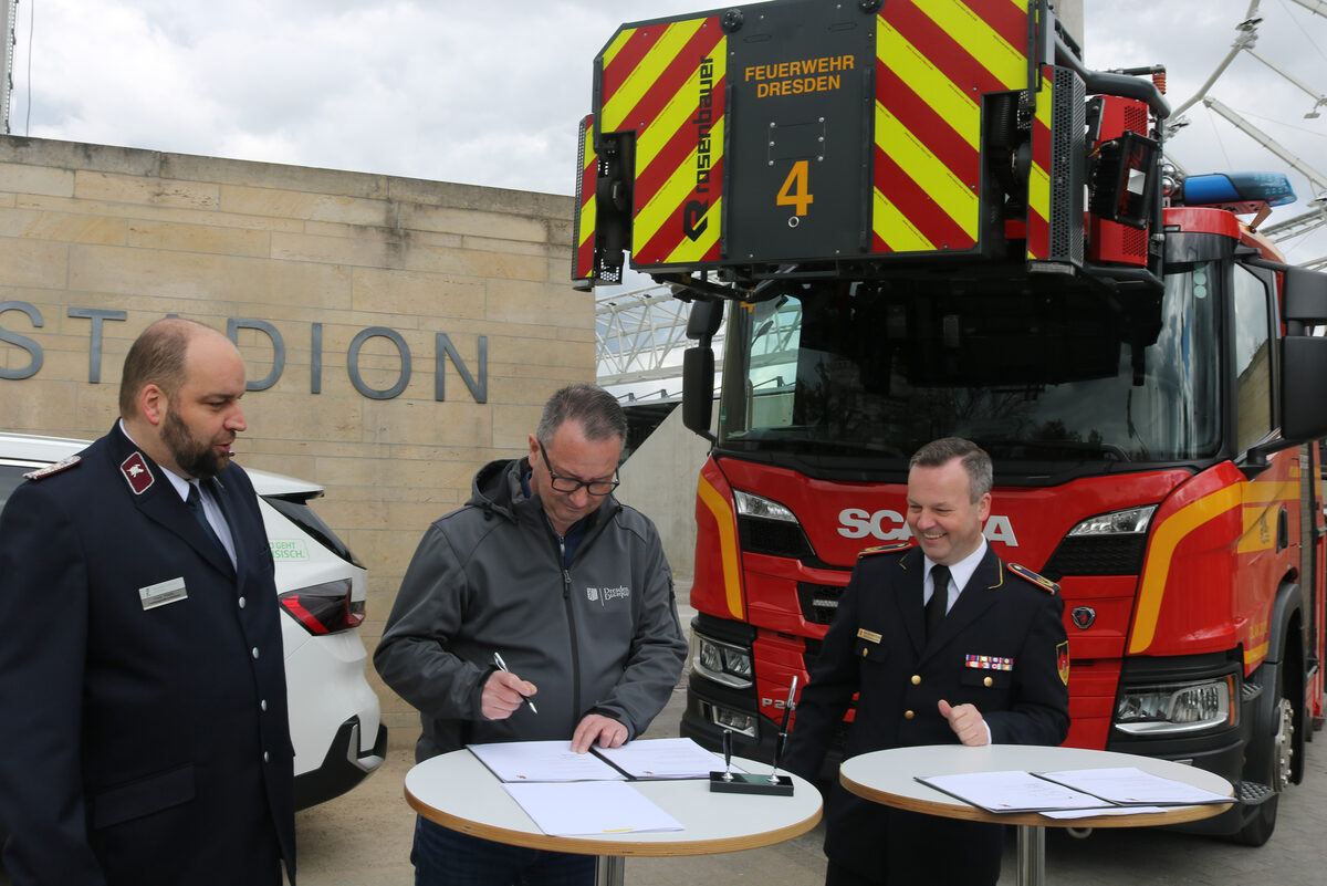 Drei Männer stehen an Tischen und unterschreiben auf Papier, im Hintergrund steht eine Feuerwehr