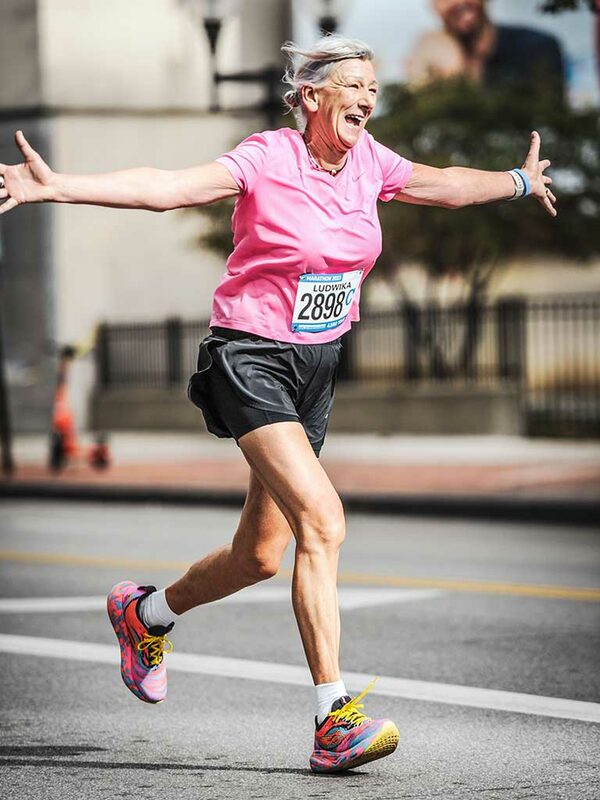 Ältere Frau mit grauen Haaren in rosa Shirt und schwarzen Shorts und Laufschuhen läuft auf einer Straße und steckt freudig die Hände zur Seite