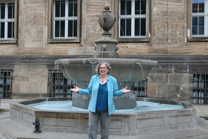 Frau steht vor einem sprudenlden Brunnen, im Hintegrund befinden sich Gebäude