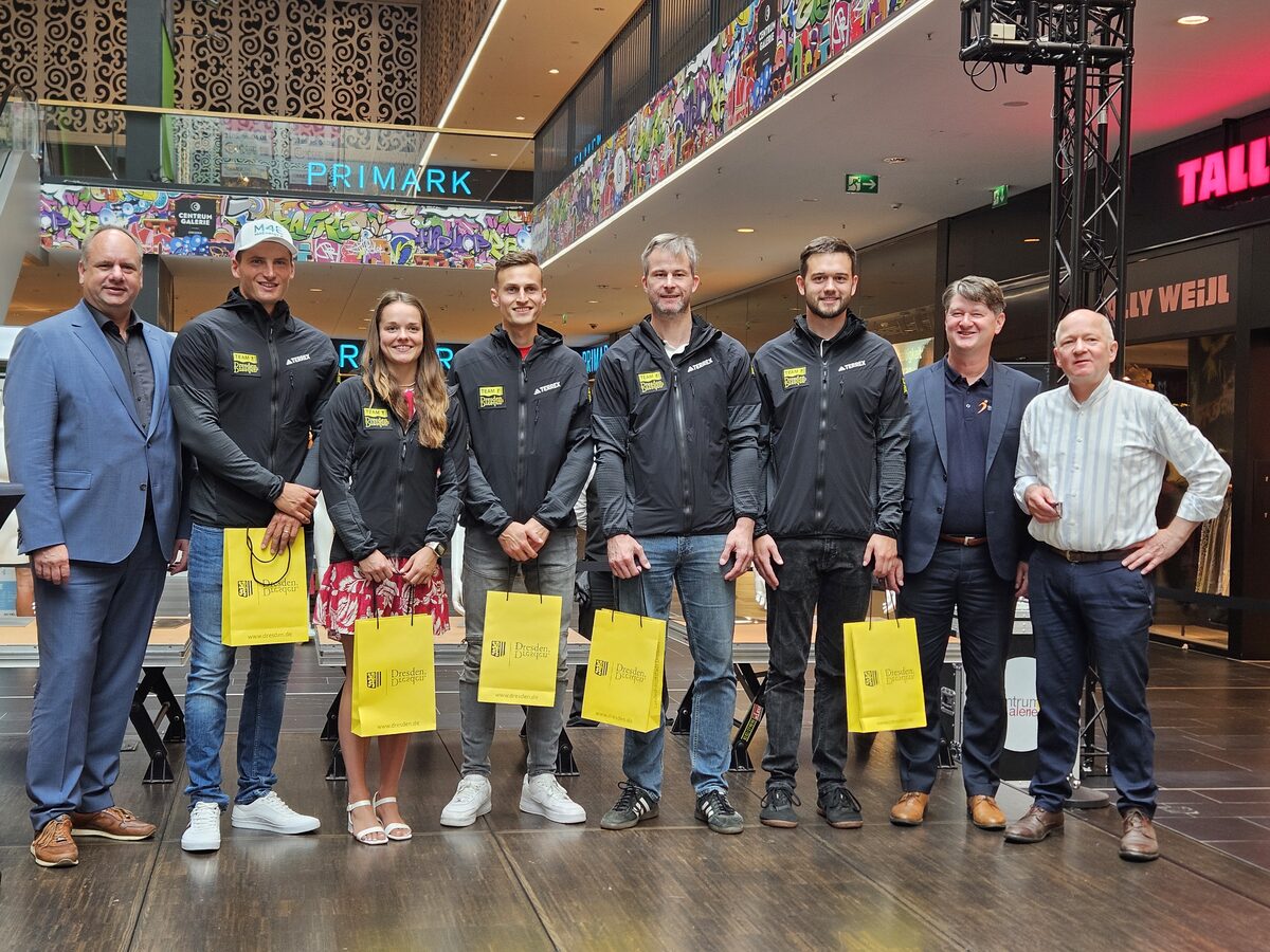 7 Männer und eine Frau in einer Reihe, davon 5 mit einer gelben Tüte in der Hand in einem Einkaufszentrum