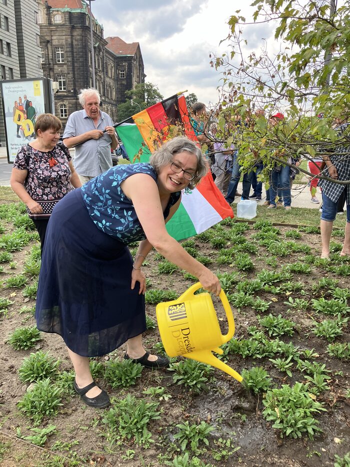 Frau steht in Blumenbeet und gießt mit einer gelben Gießkanne mit der Beschriftung "Dresden" die Pflanzen. Im Hintergrund stehen Menschen und ein Mann hält eine Flagge mit den Farben rot-schwarz-rot-gelb-grün-weiß