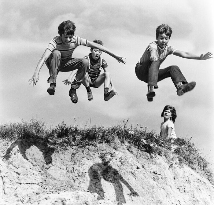 Schwarz-Weiß Foto: Drei Kinder springen über eine Düne, ein Kind guckt zu