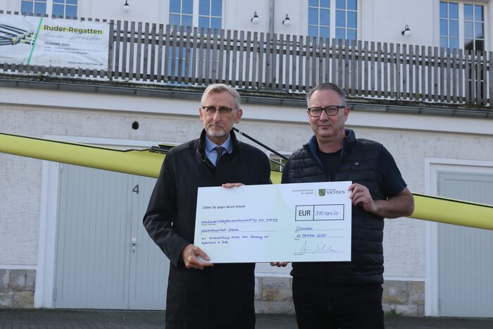 Zwei Personen halten einen Förderscheck in die Kamera und stehen vor dem Bootshaus