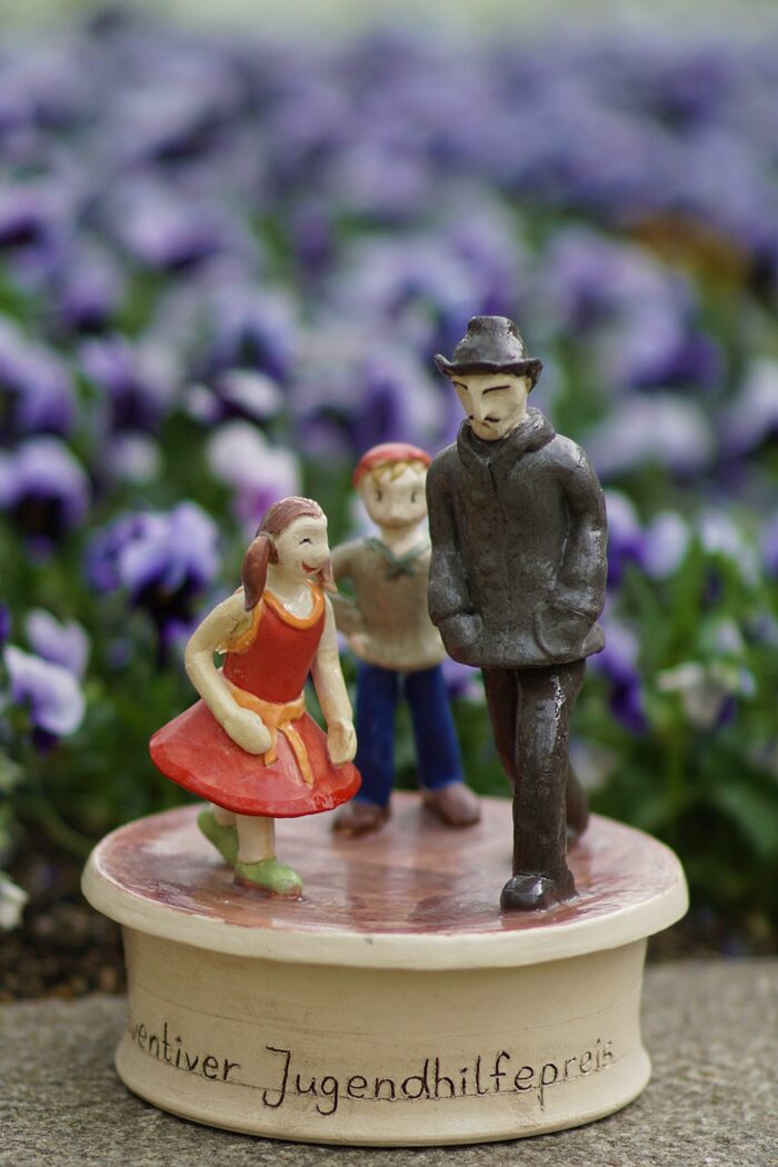 Pokal aus Keramik mit einem Mädchen im roten Kleid, einem Kind mit blauer Hose und grünem Shirt, sowie einem Mann mit schwarzem Mantel und Hut