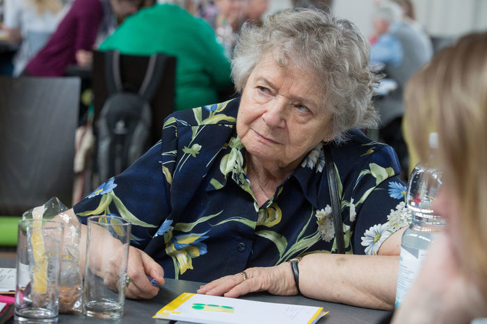 Ein ältere Frau sitzt an einem Tisch und hört interessiert zu.