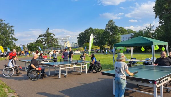 Tischtennisplatten unter freiem Himmel, an dem Menschen mit und ohne Rollstuhl spielen, im Hintergrund ein Fest
