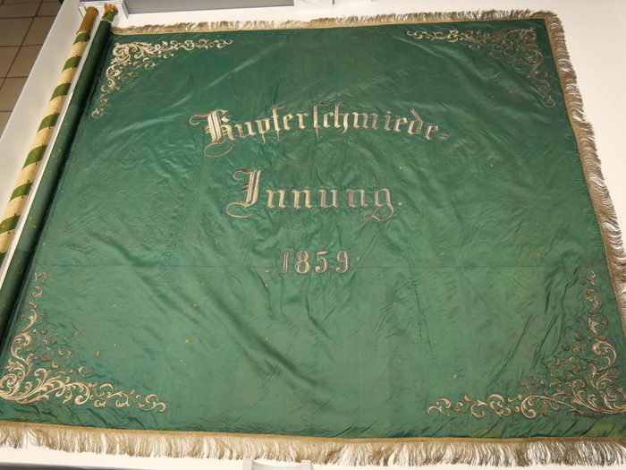Innungsfahne der Kupferschmiede aus dem Jahr 1859. Die grüne Seidenfahne zeigt florale Siedenstickereien an den Ecken und die Inschrift "Kupferschmiedeinnung 1859"