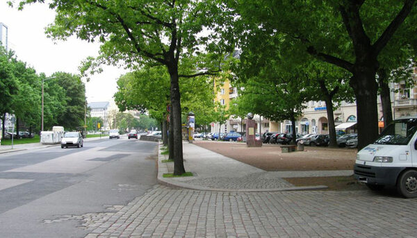 Das Bild zeigt den teilweise neu gestalteten Bönischplatz mit den an dessen Südseite gelegenen Ladengeschäften, die von überdimensionierten Straßenverkehrsflächen beeinträchtigt werden.