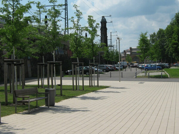 Das Bild zeigt die mit einem Baumdach gestaltete Teilfläche und im Hintergrund den mit privaten Mitteln neu gestalteten Parkplatz