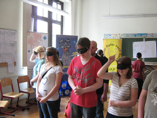 Im Foto ist zu sehen wie Schüler stehend mit verbundenen Augen vor dem Projektleiter stehen und eine Berührung mit einer Stoffkeule am Rücken zu lassen