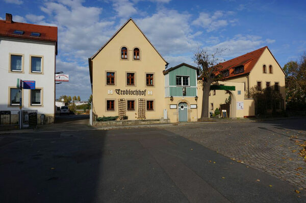 Südansicht des Trobischhofes in Alttrachau. Zu sehen sind jeweils die Giebel der beiden Gebäude, welche zur Straße ausgerichtet sind. Links neben den warmgelben Gebäuden des Trobischhofes ist ein traufständiges, weißes Haus mit roten Dachziegeln zu sehen, welches einen Kfz-Service und einen Reifendienst beherbergt.