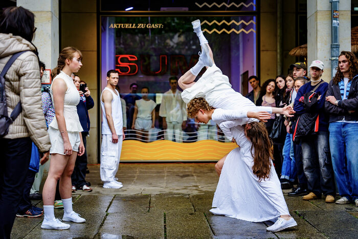 Fensterblicke aus dem Ladengeschäft der Hauptstraße 42-44 auf die Tanzgeschicke im Straßenraum. Eine Tänzerin schwingt sich über den Rücken einer anderen. Weitere weißgekleidete Tanzende und Zuschauende beobachten die Tanzfigur.
