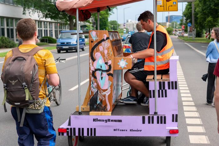 Ein Mann spielt auf einem Klavier, das auf einem selbstgebauten Anhänger von einem Radfahrer über die Albertstraße gezogen wird - begleitet Polizei und Zuhörenden.