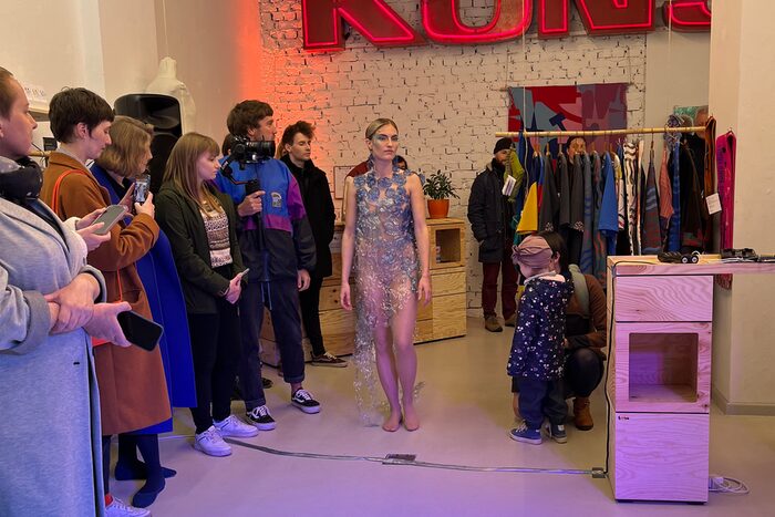 Mit einer Modenschau startete das dreiwöchigen Slow-Fashion-Festival. Ein Model führt ein transparentes silbern schimmerndes Kleid den Zuschauenden vor, die am Rand des Raumes stehen.