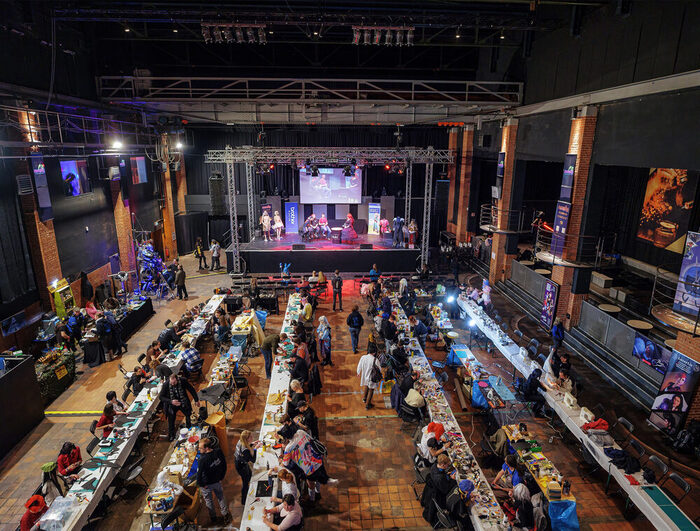 Blick von oben auf Tischreihen und eine Bühne im Stromwerk der Kulturarena Kraftwerk Mitte Dresden. An den Tischen basteln viele Menschen. Auf der Bühne sind verschiedene Cosplay-Kostüme an Puppen zu sehen.
