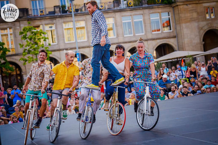 Höhepunkt des Dresdner Zirkustheater-Festival war das Fahrradspektakel der französischen "Compagnie La Bande À Tyrex" auf dem Altmarkt. Ein Mann steht auf den Lenkern zweier Fahrräder. Die beiden Radfahrenden haben sich mit drei weiteren Radelnden eingehakt und fahren gemeinsam im Kreis.