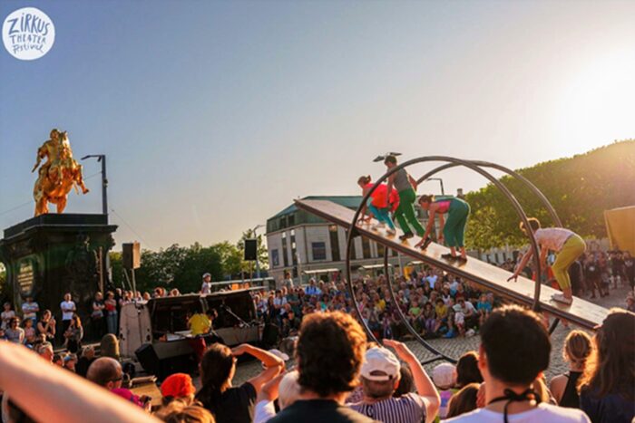 Großes Zirkus-Spektakel auf dem Neustädter Markt: Gegenüber dem "Goldenden Reiter" balancieren vier Personen ein steil nach oben ragendes Brett hoch, das in ein Rhönrad eingespannt ist.