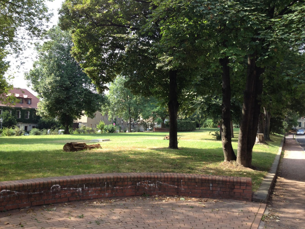 Auf dem Foto ist ein anderer Teil des Spielplatzes zu sehen, welcher mit einer kleinen Mauer eingefaßt ist. Dahinter stehen hohe Bäume auf dem grünen Rasen.