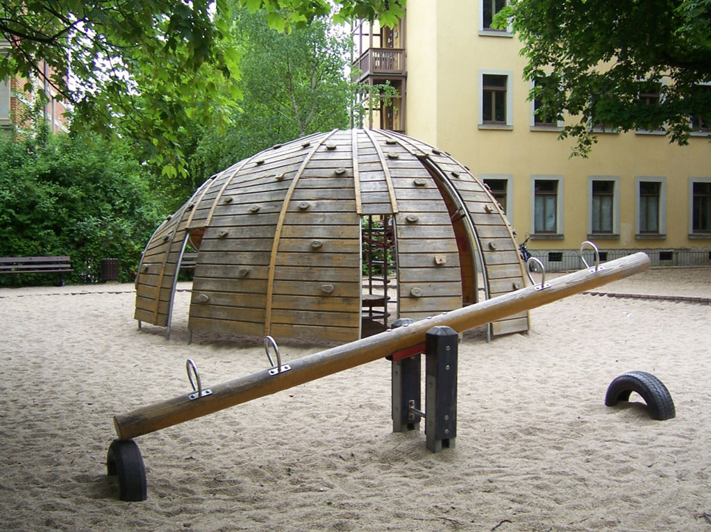 Das Foto zeigt einen Spielplatz für größere Kinder. Es ist eine halbrunde Holzkugel, welche mit Klettergriffen versehen ist, eine Wippe und Sitzbänke für Eltern.