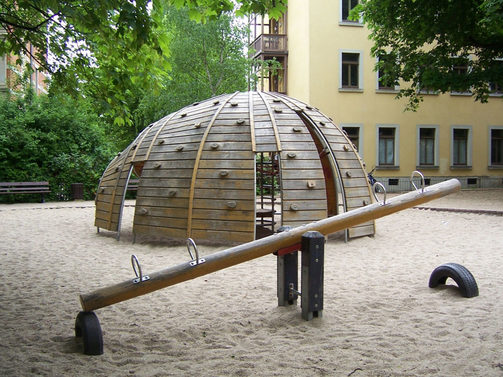 Das Foto zeigt einen Spielplatz für größere Kinder. Es ist eine halbrunde Holzkugel, welche mit Klettergriffen versehen ist. Außerdem sind eine Wippe und Sitzbänke für Eltern zu sehen.