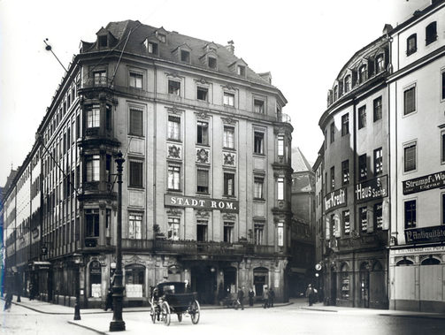 Historischer Standort des "Hotel Stadt Rom" am Neumarkt. Platsraum mit Kopfsteinpflaster und Bäume im Vordergrund. 7-stöckige Wohnhäuser im Hintergrund.
