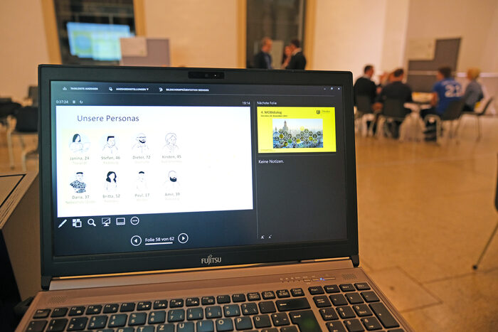 Das Foto zeigt im Vordergrund einen aufgeklappten Laptop mit den aufgeführten acht Personas sowie im Hintergrund eine Arbeitsgruppe im Gespräch.
