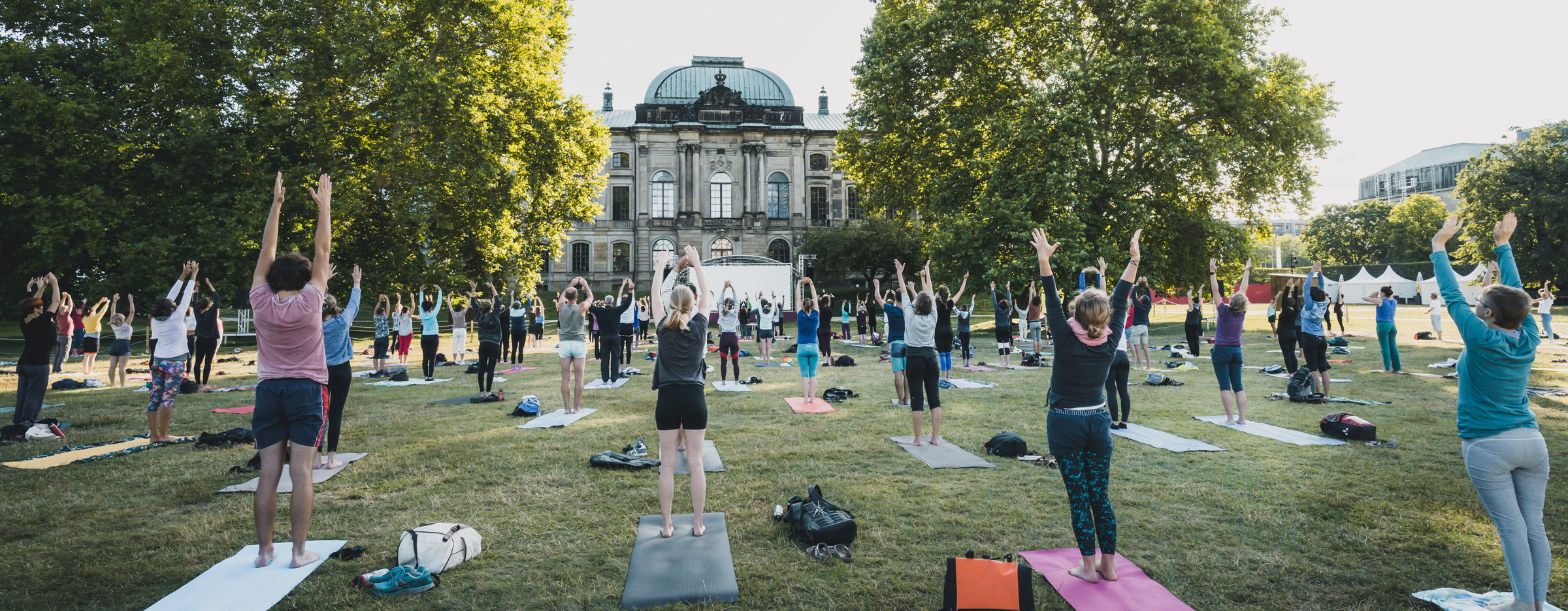 Eine große Gruppe von Frauen stehen auf Yogamatten vor dem Japanischen Palais im Palaisgarten und machen Yoga.