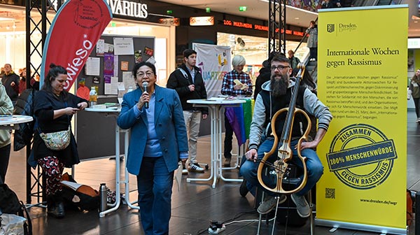 Frau in blauem Anzug neben einem Musiker daneben ein gelber Aufsteller