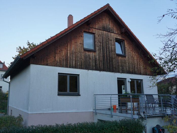 Zweigeschossiges weißes Einfamilienhaus mit Holzfassade im Giebelbereich und rotem Satteldach.