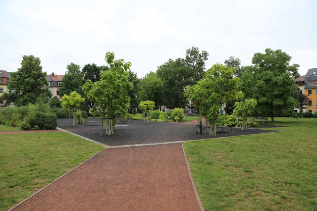 Zu sehen ist ein gestalteter Kiesplatz mit Sitzgelegenheiten und jungen Bäumen. Er ist umgeben von einer grünen Rasenfläche. Im Hintergrund erkennt man Wohnhäuser.
