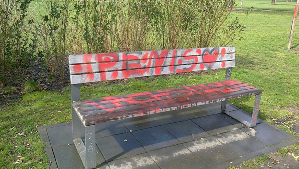 Zu sehen ist eine nasse Sitzbank, die auf der Sitzfläche und der Rückenlehne mit roten Graffiti besprüht wurde. Im Hintergrund befindet sich eine Parkanlage.