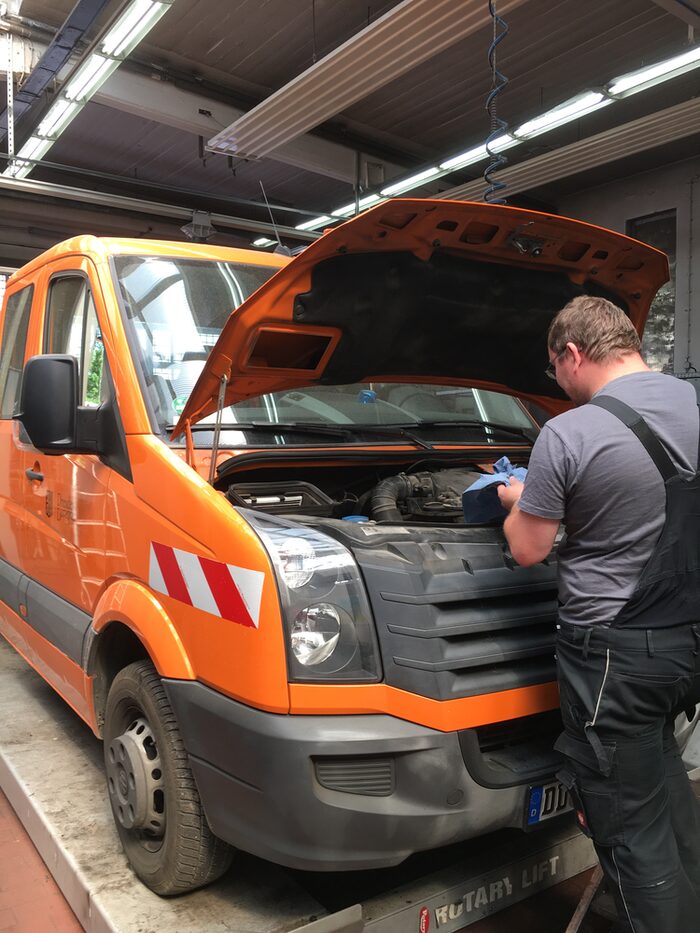 Ein Mitarbeiter in dunkler Latzhose steht mit dem Rücken zum Betrachter vor einem orangefarbenen Pritschenwagen. Die Motorhaube ist aufgeklappt, um Reparaturarbeiten vorzunehmen.