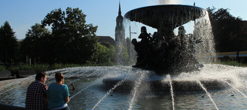 Zwei Besucher blicken auf das herausströmende Wasser des Brunnens am Albertplatz in der Neustadt. Der Himmel ist strahlend blau und wolkenlos.