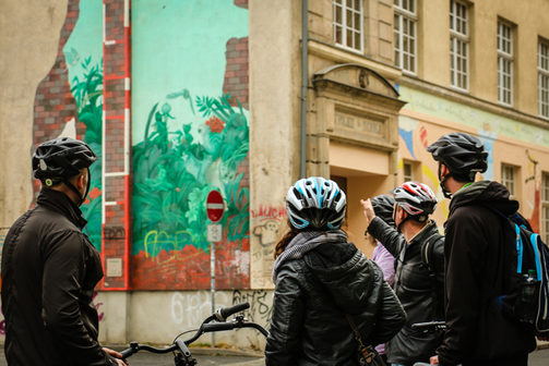 Eine Gruppe Radfahrer steht mit ihren Rädern und Helmen auf dem Kopf auf einem Gehweg. Sie schauen alle auf eine bunte Hauswand im Szeneviertel Neustadt, auf die der Gästeführer zeigt.