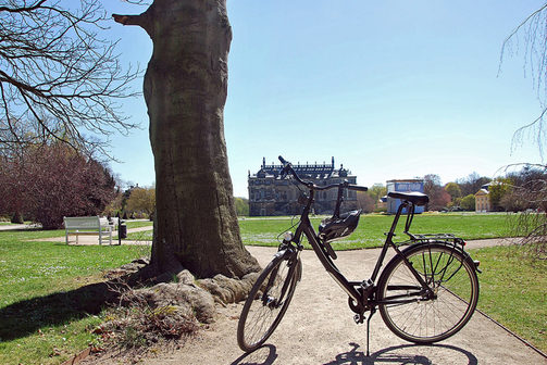 Ein Fahrrad mit Helm steht auf einem Gehweg mitten im Großen Garten. Im Hintergrund sind die grüne Wiese, eine weiße Bank sowie das Palais zu erkennen.