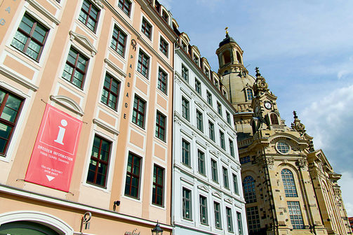 Froschperspektive auf das Quartier Frauenkirche mit einem Infobanner der Dresden Information. Daneben erstrahlt die Frauenkirche in ihrer ganzen Pracht. Der Himmel ist leicht bewölkt.