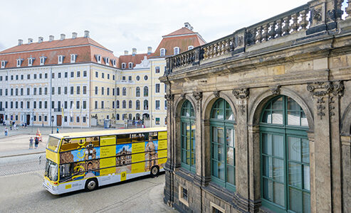 Gelber Doppeldeckerbus mit Außenwerbung und Dresden-Bildern vor dem Dresdner Zwinger. Im Hintergrund befindet sich das Taschenbergpalais.