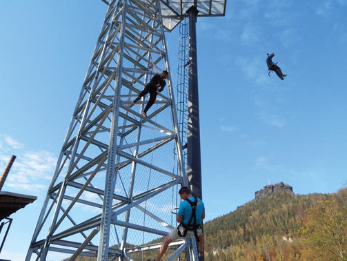 Kletterturm inmitten des Bildes. Zwei Männer befinden sich am Turm, die beide Sicherheitsgurte anhaben. Ein dritter ist im Hintergrund zu sehen, der gerade im "Flying Fox" über den Park gleitet.
