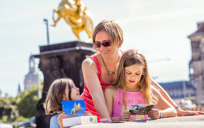 Zwei Mädchen und eine Frau stehen angelehnt am Brunnen auf der Hauptstraße mit der Statue des Goldenen Reiters und der Frauenkirche im Hintergrund. Das blonde Mädchen schaut sich eine Karte von Dresden an und das brünette Mädchen zeigt eine Karte mit Motiv des Goldenen Reiters nach oben.