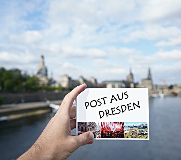 Postkarte wird von einer Hand gehalten. Im Hintergrund sieht man verschwommen die Brühlsche Terrasse