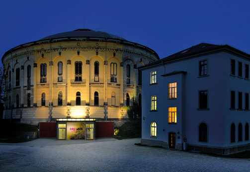 Die Außenansicht des Panometers am Abend bei wolkenlosen Himmel. Auf den Scheiben der Eingangstür steht in Großbuchstaben Dresden.