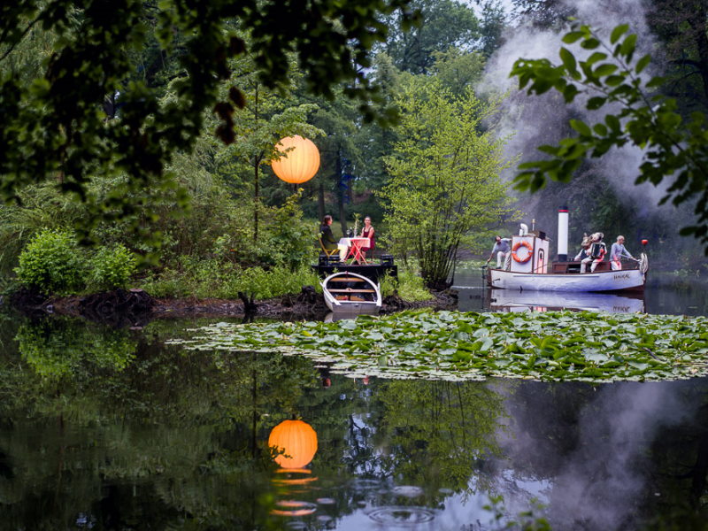 Boote auf einem Teich mit vielen Seerosen. Ein Pärchen sitzt an einem gedeckten Tisch auf einer kleinen Insel und über ihnen baumelt ein orangefarbener Lampion.