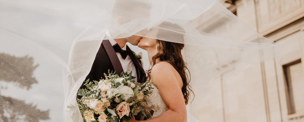 Hochzeitspaar küsst sich unter dem großen, weißen Schleier. Die Braut hält den Brautstrauß in der Hand, der im Vordergrund des Bildes zu sehen ist.